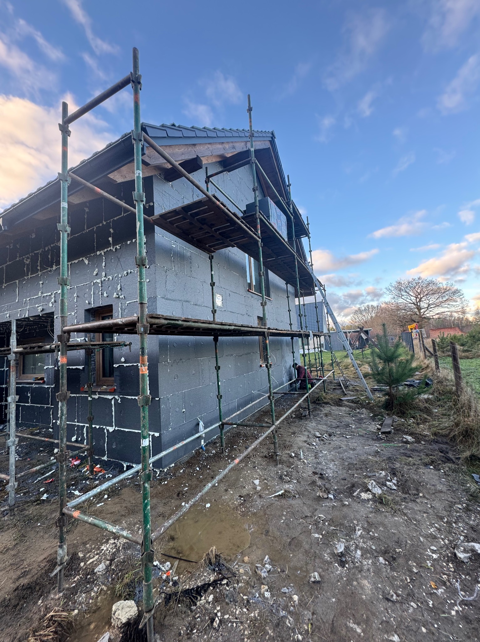 Dom w trakcie ocieplania, widoczne rusztowania i pracownik przy elewacji. Szare płyty izolacyjne z białymi elementami mocującymi. Tło: niebo i drzewa.