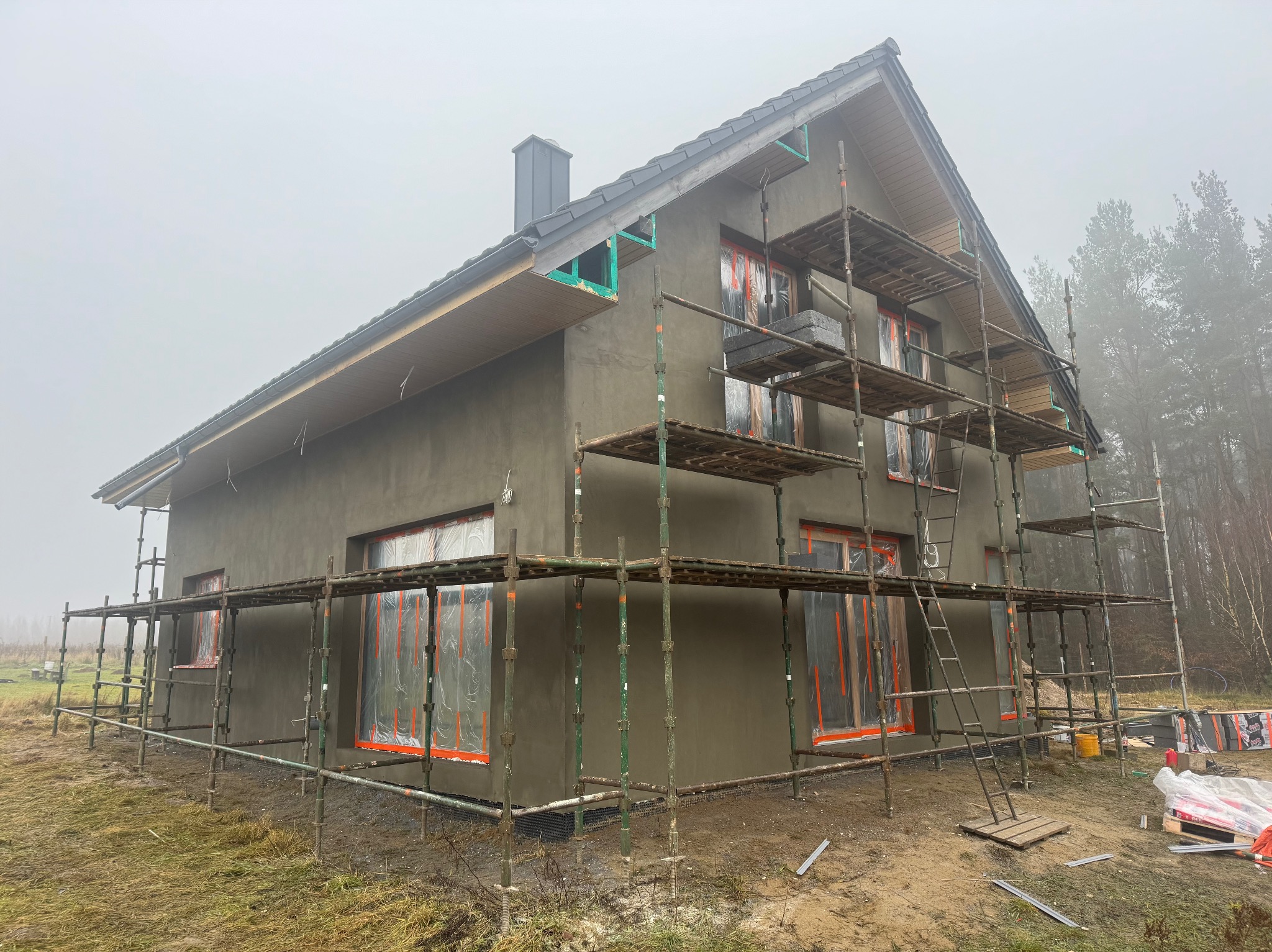 Dom jednorodzinny w trakcie nakładania szarego tynku elewacyjnego, z rusztowaniem i zabezpieczonymi oknami folią. Widok na budynek w pochmurny dzień.