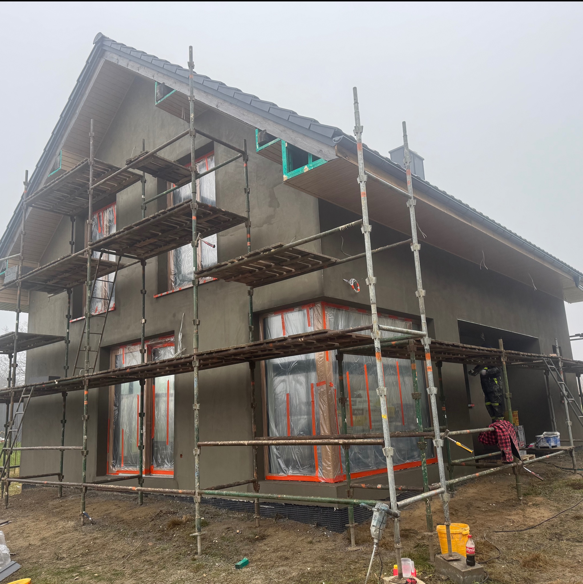 Dom w trakcie nakładania szarego tynku elewacyjnego, z rusztowaniami i oknami zabezpieczonymi folią. Widoczny fragment dachu i otoczenie placu budowy.