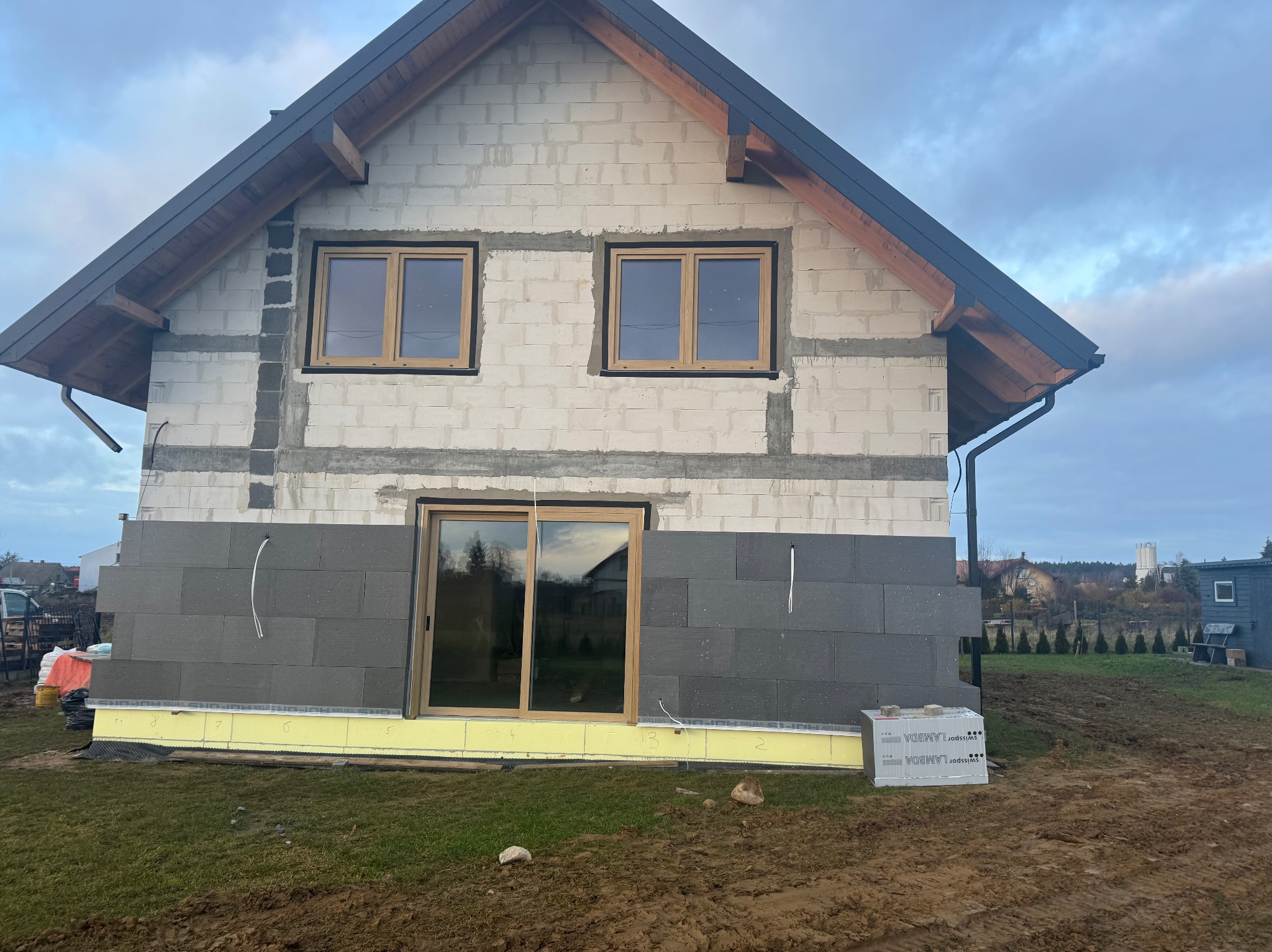 Dom w trakcie ocieplania: szare płyty izolacyjne na dolnej części ściany, okna z drewnianymi ramami, ściany z białego bloczka. Widoczne materiały izolacyjne.