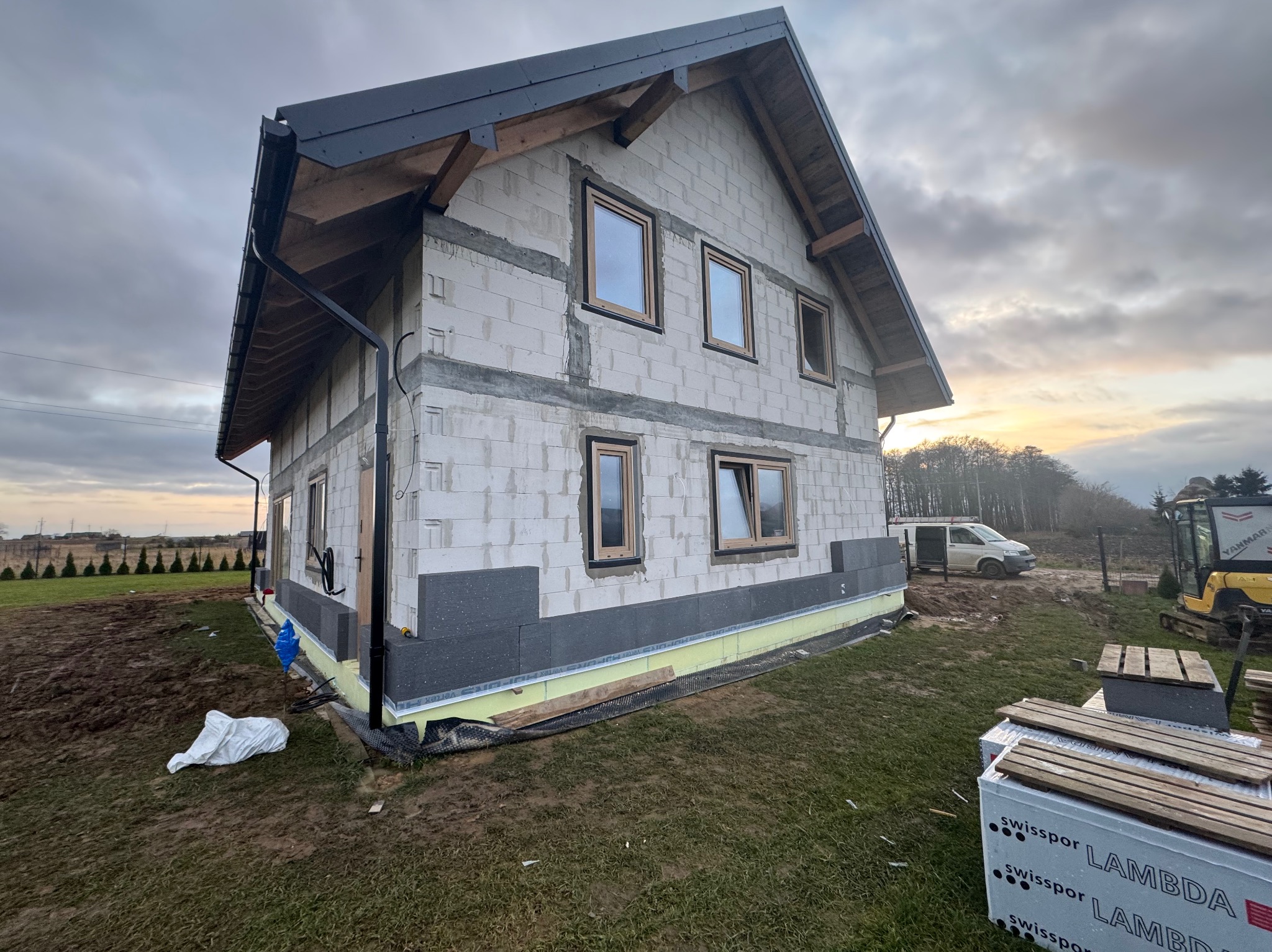 Surowy dom z białego bloczka, częściowo ocieplony szarym styropianem, z zamontowanymi oknami. Widoczne przygotowania do dalszych prac budowlanych.