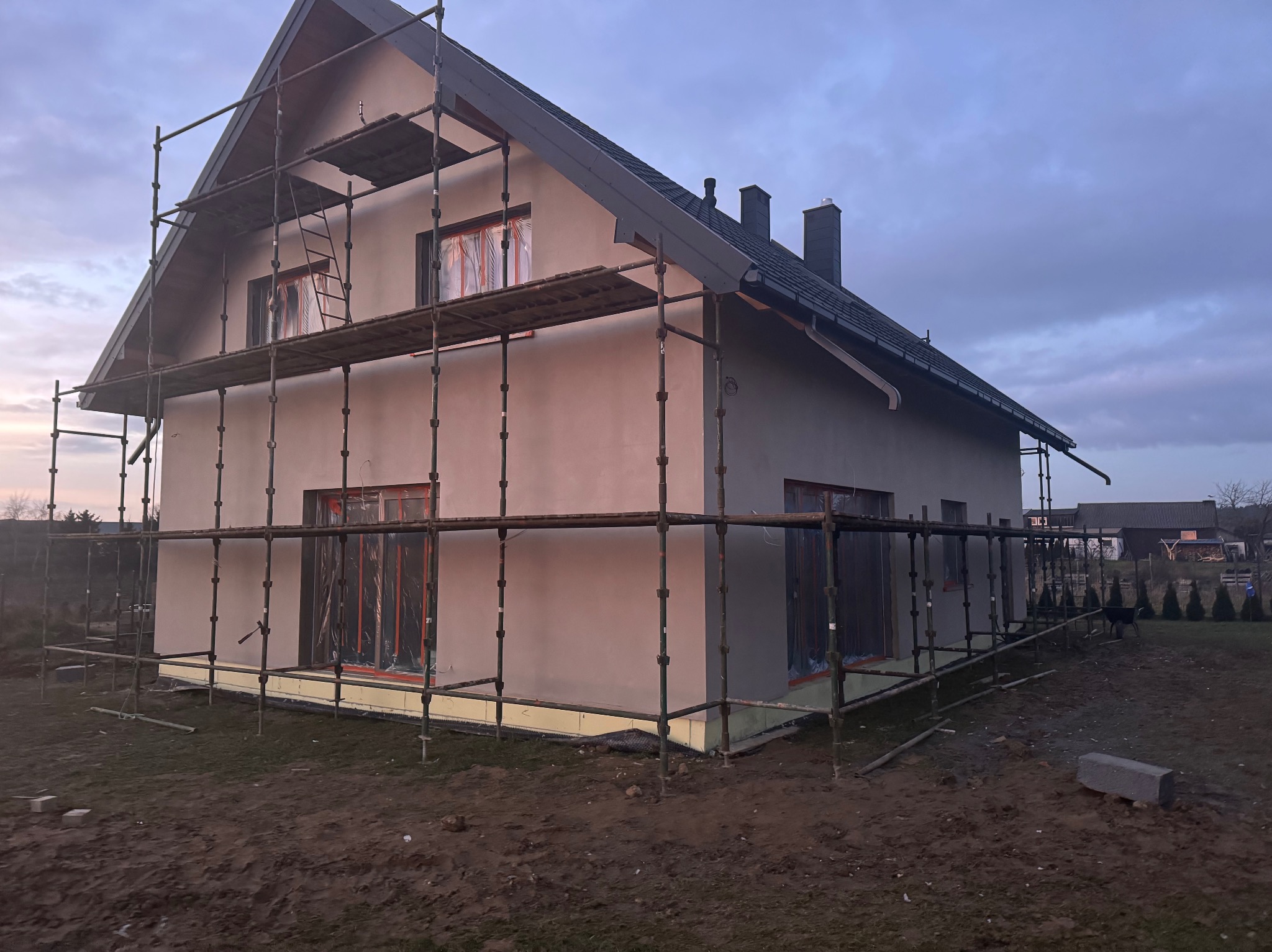 Dom jednorodzinny w trakcie prac elewacyjnych, z widocznymi rusztowaniami i zabezpieczonymi oknami. Świeżo nałożony tynk w odcieniach szarości.