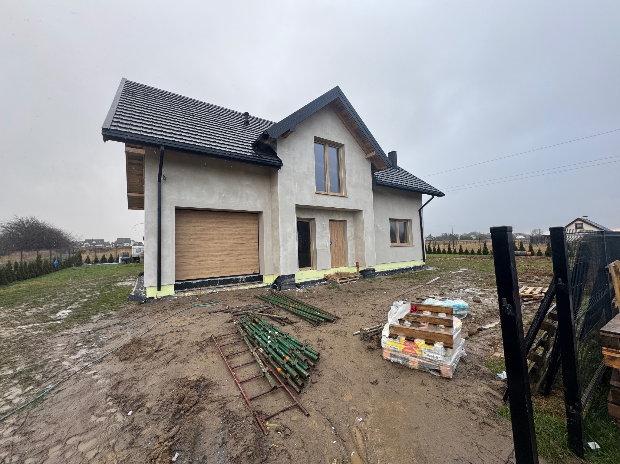 Surowy dom jednorodzinny z szarą elewacją i drewnianymi elementami wykończeniowymi, widoczny w pochmurny dzień. Na pierwszym planie materiały budowlane i błoto.