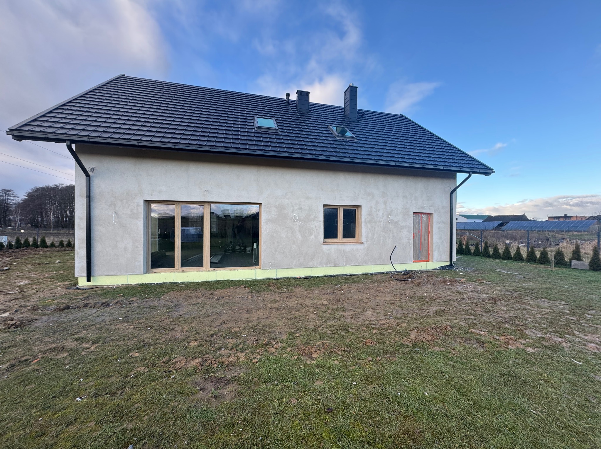 Niedokończony dom jednorodzinny z ciemną dachówką, otynkowany, z widocznymi oknami i drzwiami. W tle błękitne niebo i fragment ogrodu z tujami.