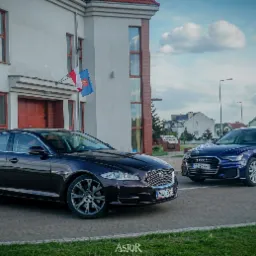Dwa luksusowe samochody, Jaguar w kolorze burgundowym i Audi w kolorze granatowym, udekorowane ślubnymi ozdobami, zaparkowane przed budynkiem z polską flagą.