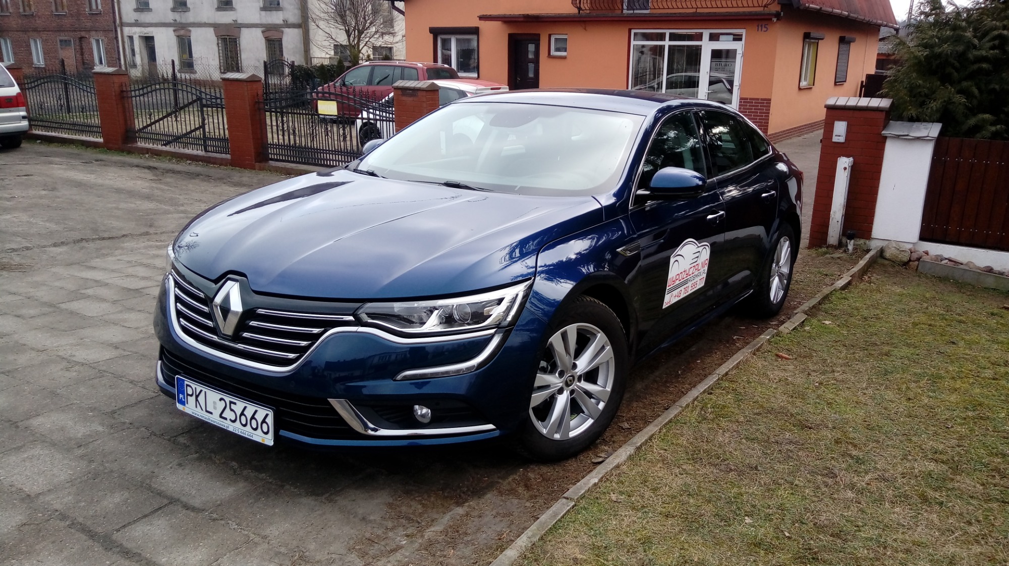 Granatowy samochód Renault Talisman z naklejką firmy na boku, zaparkowany na podjeździe przed budynkiem.