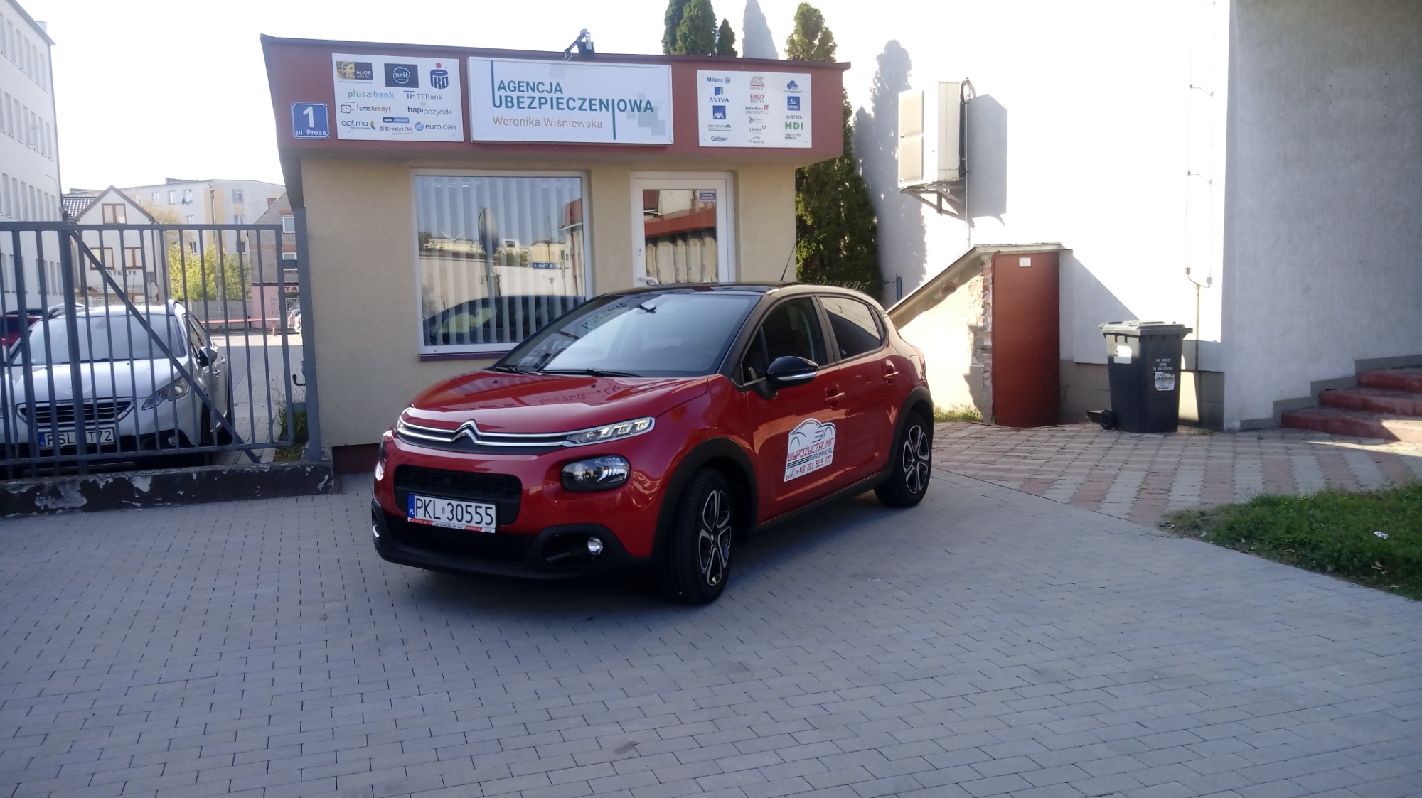 Czerwony samochód osobowy z naklejką 'Wypożyczalnia' zaparkowany przed budynkiem agencji ubezpieczeniowej z widocznymi szyldami różnych firm finansowych.