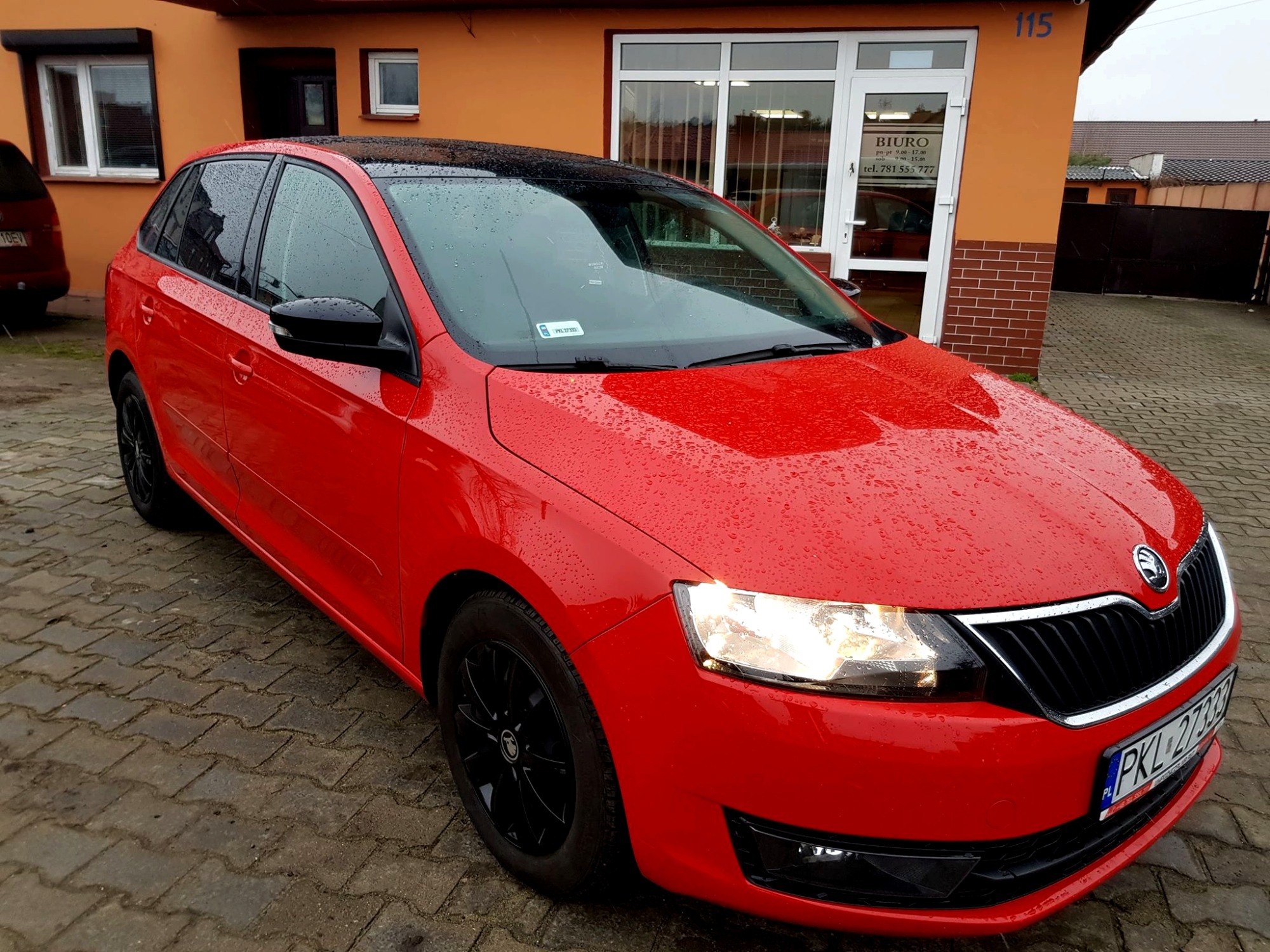 Czerwony samochód Skoda Rapid z czarnym dachem i felgami, zaparkowany na brukowanej nawierzchni przed budynkiem z szyldem biura.