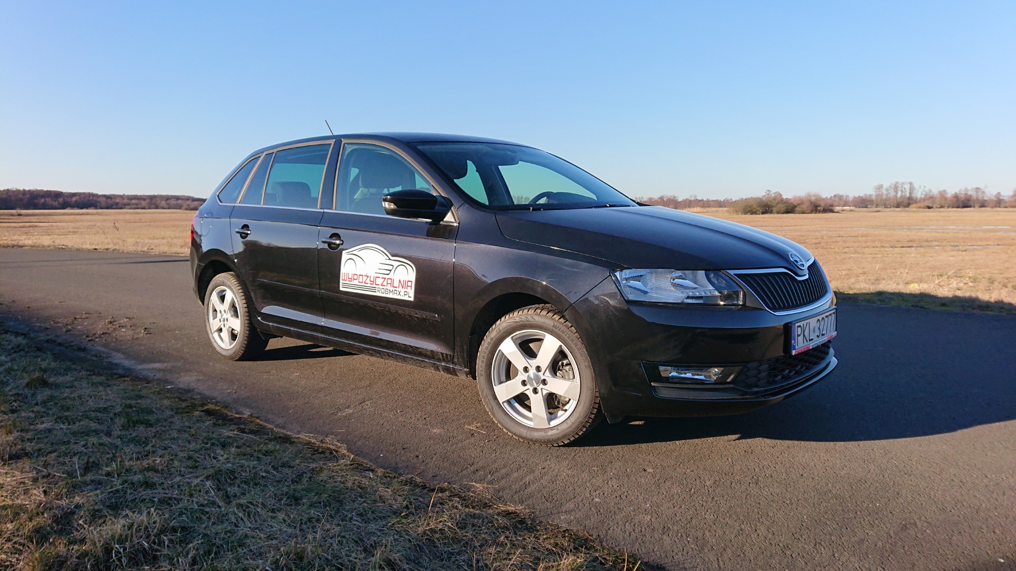 Czarna Skoda Rapid na asfalcie z naklejką firmy na drzwiach, w tle pole i błękitne niebo.