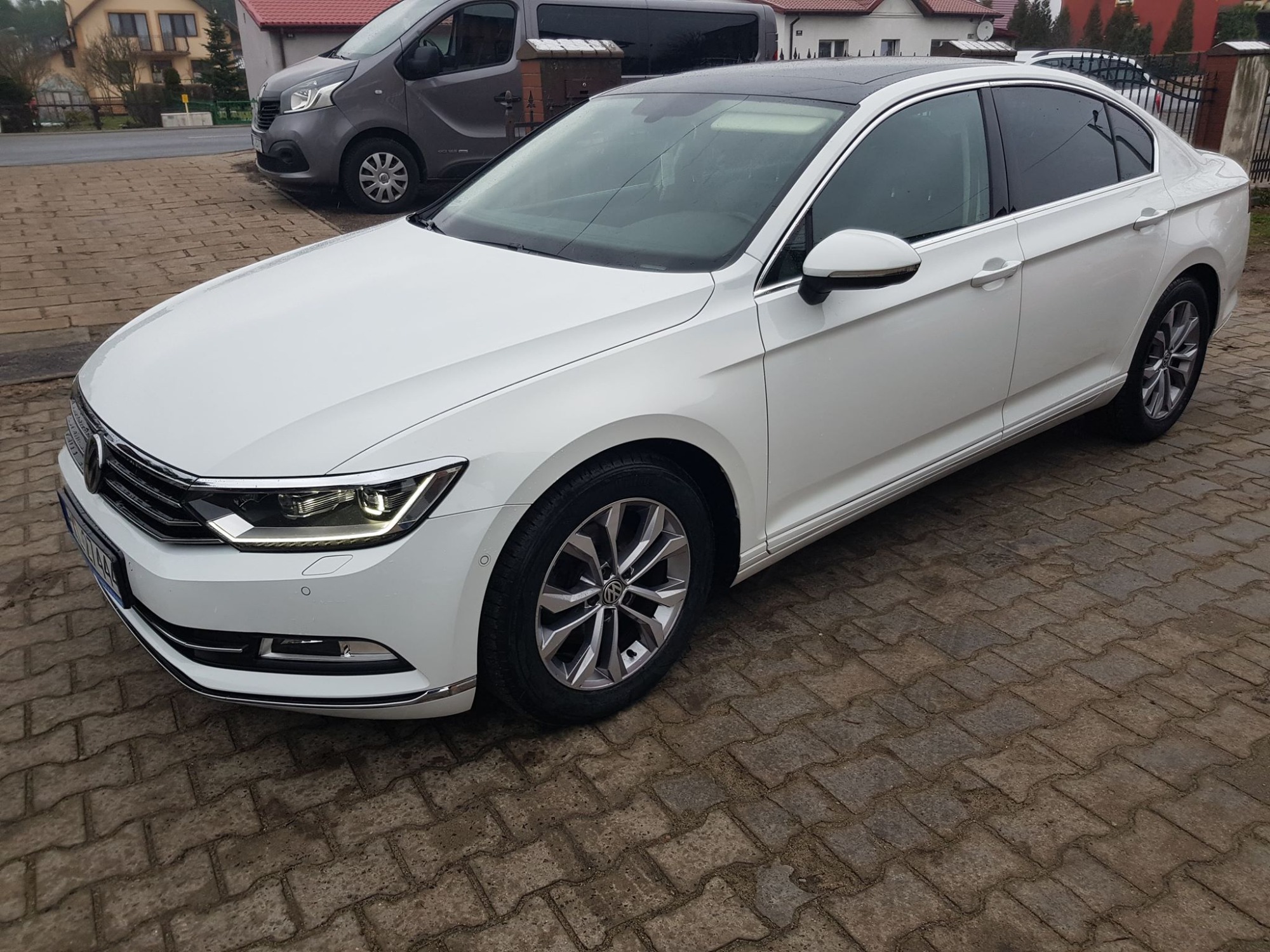 Biały sedan z czarnym dachem, zaparkowany na kostce brukowej, z widocznym fragmentem szarego vana i zabudowań w tle.