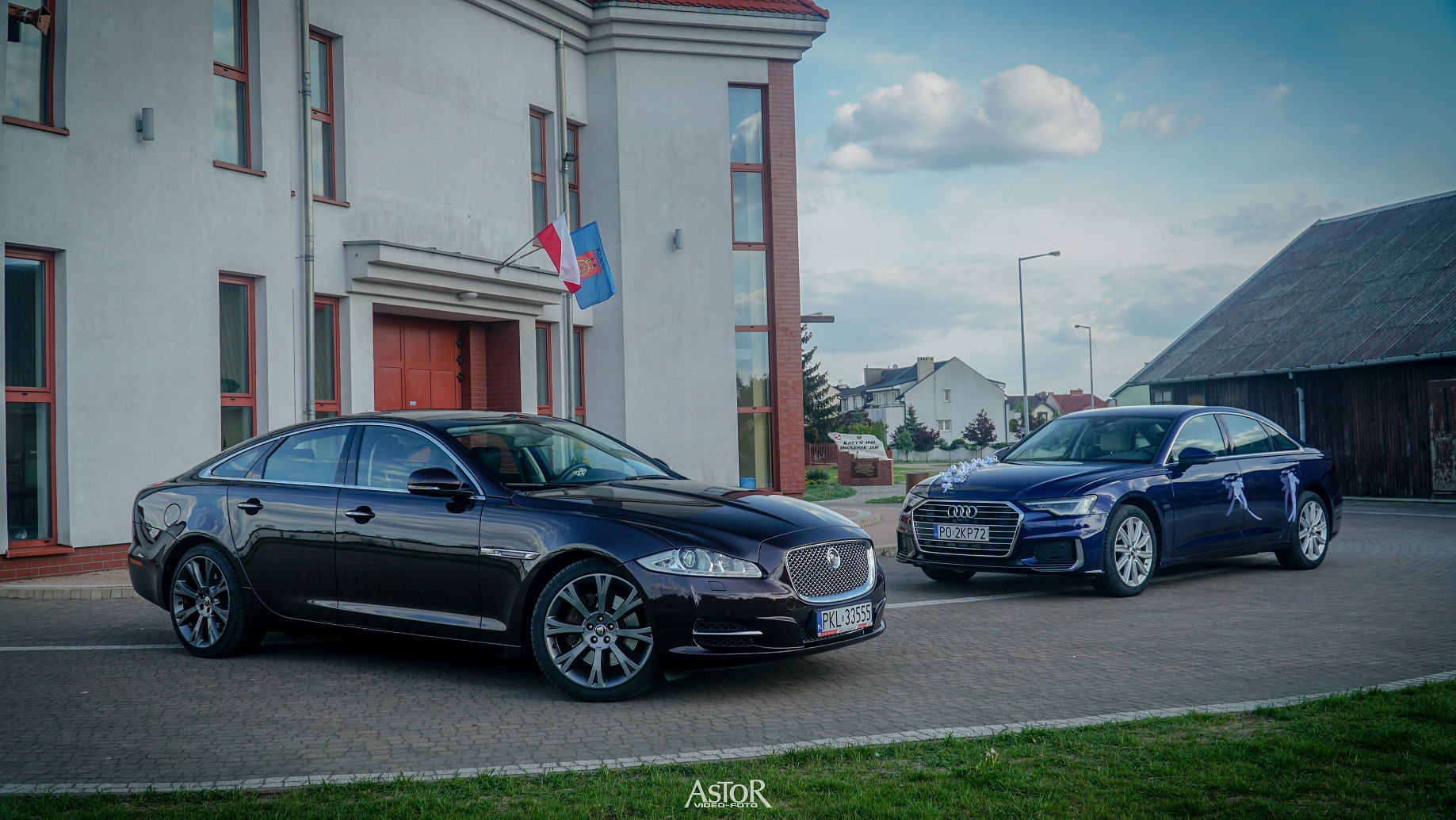 Dwa luksusowe samochody, Jaguar w kolorze burgundowym i Audi w kolorze granatowym, udekorowane ślubnymi ozdobami, zaparkowane przed budynkiem z polską flagą.