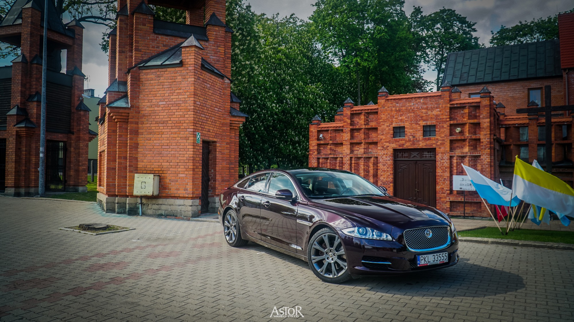 Bordowy Jaguar XJ zaparkowany na brukowanym placu przed budynkiem z czerwonej cegły z flagami w tle.