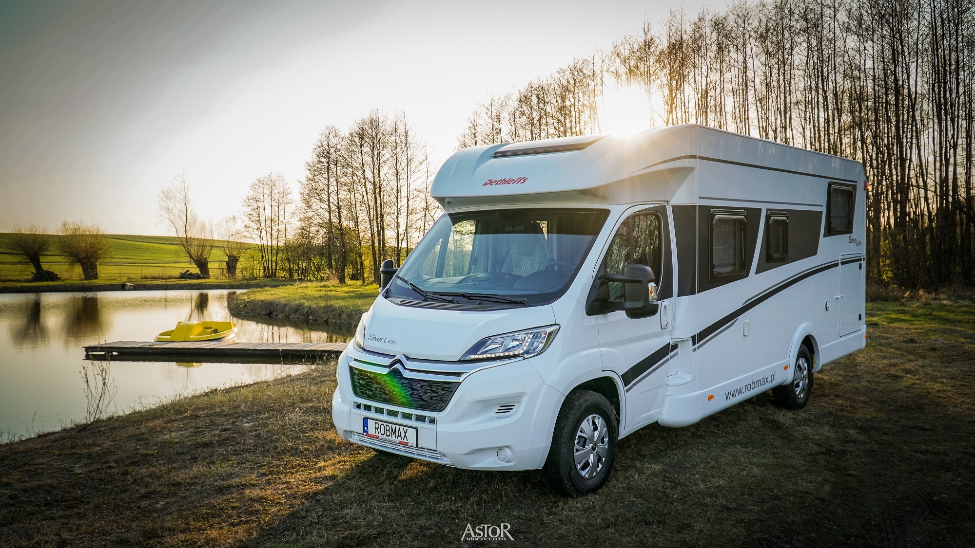 Biały kamper Dethleffs Silver Line na trawiastym brzegu jeziora z żółtym rowerem wodnym na pomoście, w tle las i zachodzące słońce.