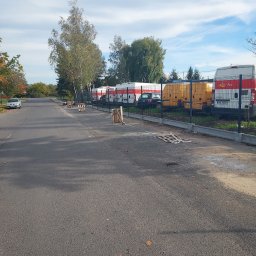 Usługi Ogólnobudowlane Marian Lasyk - Ulica z nowym betonowym obrzeżem chodnika, za którym znajduje się ogrodzenie z siatki i zaparkowane pojazdy pocztowe.