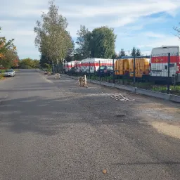 Ulica z nowym betonowym obrzeżem chodnika, za kt&oacute;rym znajduje się ogrodzenie z siatki i zaparkowane pojazdy pocztowe.