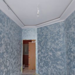 Usługi Ogólnobudowlane Marian Lasyk - Ściany w pokoju po dekoracyjnym malowaniu w odcieniach szarości i błękitu, widoczny sufit z listwami i niedokończone oświetlenie, na podłodze narzędzia malarskie i taśmy zabezpieczające.