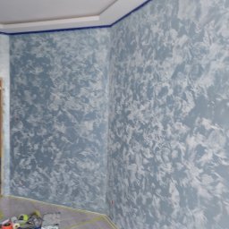 Usługi Ogólnobudowlane Marian Lasyk - Pokój po malowaniu ścian na szaro-biało z efektem dekoracyjnym, zabezpieczona taśmą listwa sufitowa w kolorze granatowym, podłoga zabezpieczona folią, widoczne narzędzia malarskie.