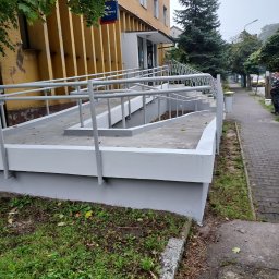 Usługi Ogólnobudowlane Marian Lasyk - Zewnętrzna rampa dla niepełnosprawnych, pomalowana na szaro, prowadząca do budynku poczty, widok z poziomu chodnika.