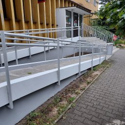 Usługi Ogólnobudowlane Marian Lasyk - Rampa dla niepełnosprawnych prowadząca do wejścia Poczty Bankowej, świeżo pomalowana na szaro, z widocznym fragmentem elewacji budynku w odcieniach żółci i brązu.