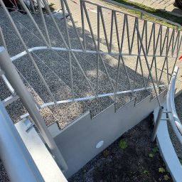 Usługi Ogólnobudowlane Marian Lasyk - Zewnętrzne schody z szarym, kamyczkowym wykończeniem stopni i szarą, metalową balustradą o geometrycznym wzorze, widoczne z góry pod kątem.