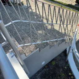 Zewnętrzne schody z szarym, kamyczkowym wykończeniem stopni i szarą, metalową balustradą o geometrycznym wzorze, widoczne z góry pod kątem.