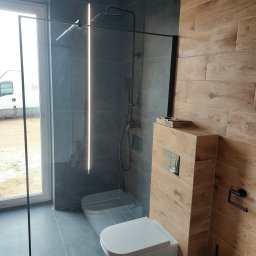 Usługi Ogólnobudowlane Marian Lasyk - Nowoczesna łazienka z kabiną prysznicową typu walk-in z czarnymi profilami, podwieszaną toaletą i drewnopodobnymi płytkami na ścianie, z widoczną linią ledową w kabinie.