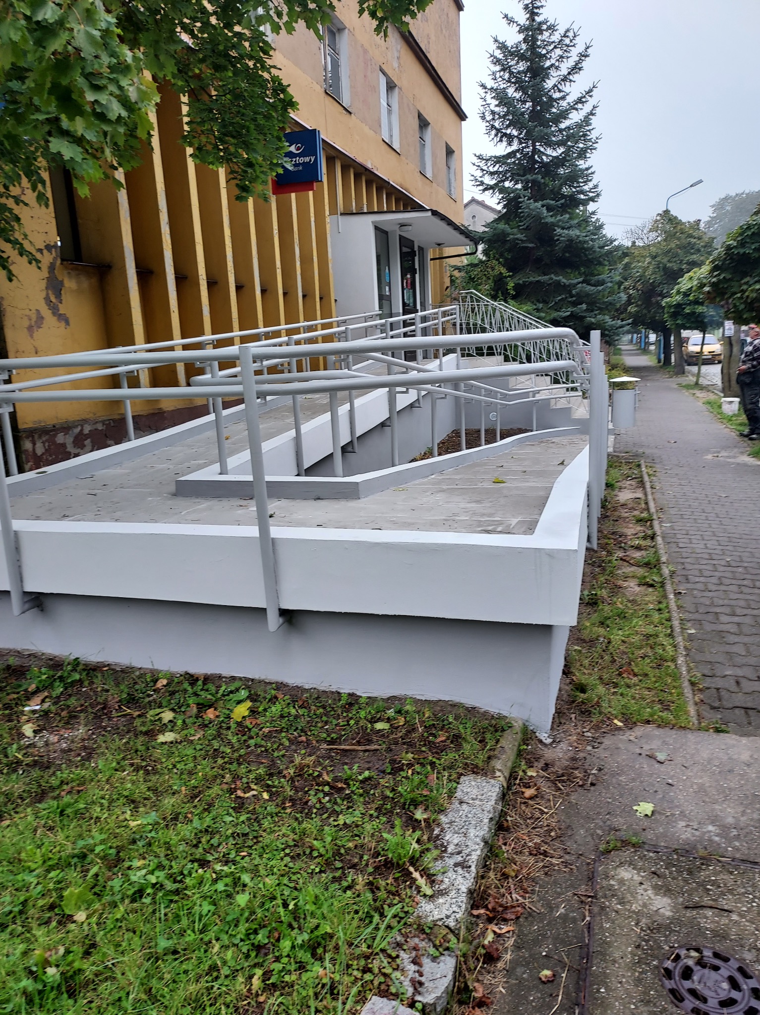 Zewnętrzna rampa dla niepełnosprawnych, pomalowana na szaro, prowadząca do budynku poczty, widok z poziomu chodnika.