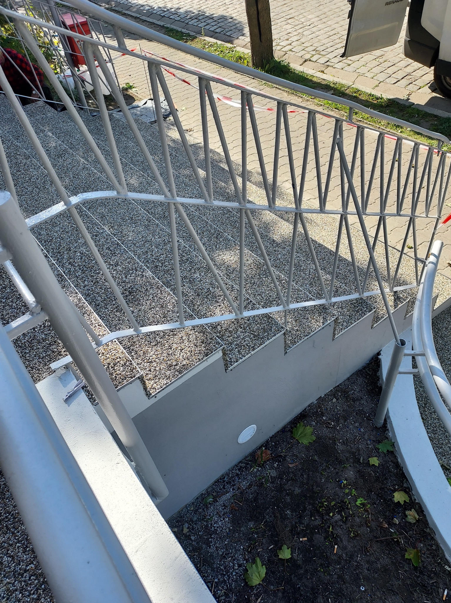 Zewnętrzne schody z szarym, kamyczkowym wykończeniem stopni i szarą, metalową balustradą o geometrycznym wzorze, widoczne z góry pod kątem.