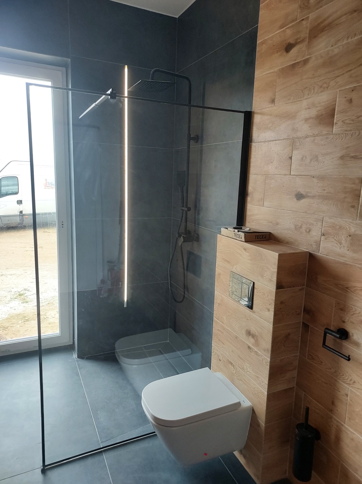 Nowoczesna łazienka z kabiną prysznicową typu walk-in z czarnymi profilami, podwieszaną toaletą i drewnopodobnymi płytkami na ścianie, z widoczną linią ledową w kabinie.