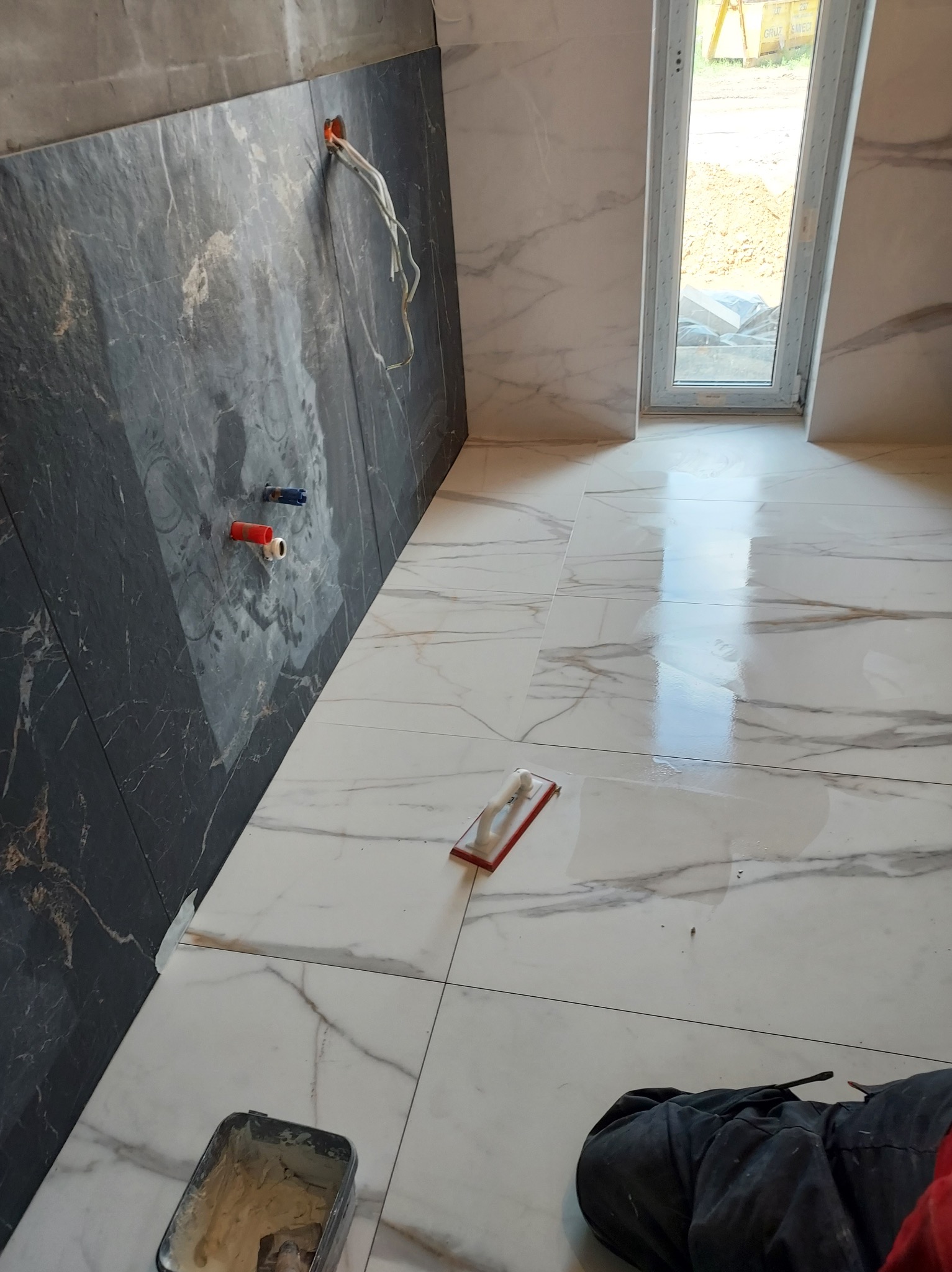 Układanie białych płytek imitujących marmur na podłodze i czarnych na ścianie, widoczne wystające kable i rury, fugowanie w trakcie, narzędzia pracy na podłodze.
