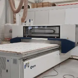 Duża frezarka CNC z białym korpusem i niebieskimi elementami, obrabiająca płytę z zaznaczonym rastrem, w tle widoczne rury odciągowe i ściany hali.