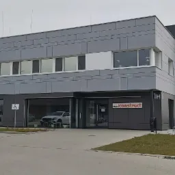 Nowoczesny, dwukondygnacyjny budynek biurowy z szarymi panelami elewacyjnymi, widoczny biały samochód osobowy na parkingu oraz logo firmy 'expo construct' nad wejściem.