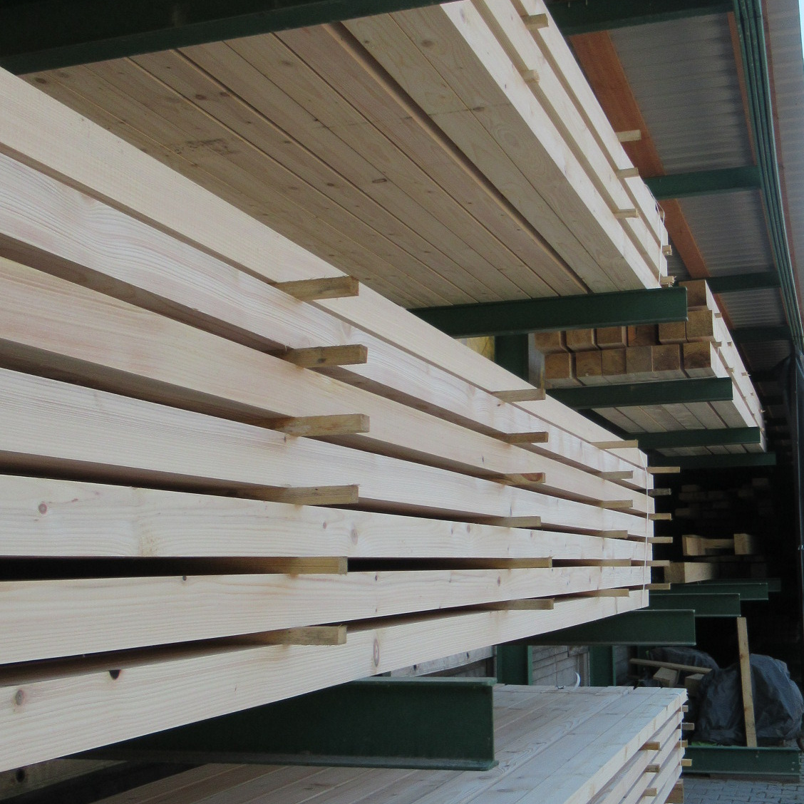 Stosy jasnej, surowej kantówki ułożone na metalowych regałach w magazynie, widoczne sęki i usłojenie drewna.