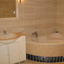 Jasna łazienka z narożną wanną obudowaną mozaiką i szafką z umywalką, wykończona beżowymi płytkami z dekoracyjnym pasem.