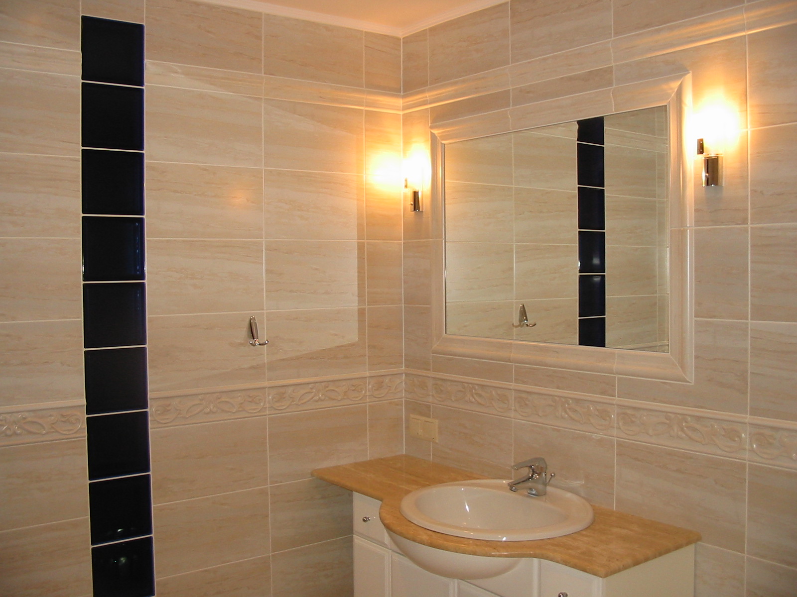 Beżowa łazienka z dekoracyjną kolumną z ciemnych płytek, owalną umywalką na blacie z jasnego marmuru i lustrem w białej ramie, oświetlona kinkietami.