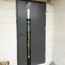 Montaż szarych drzwi zewnętrznych z pionowym przeszkleniem w surowym murze z betonu komórkowego; widoczna czerwona klamka i narzędzia w kartonie, etap prac wykończeniowych.