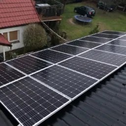 Panel słoneczny na dachu domu jednorodzinnego, widok z góry, na tle trawnik z trampoliną i dwoma samochodami.