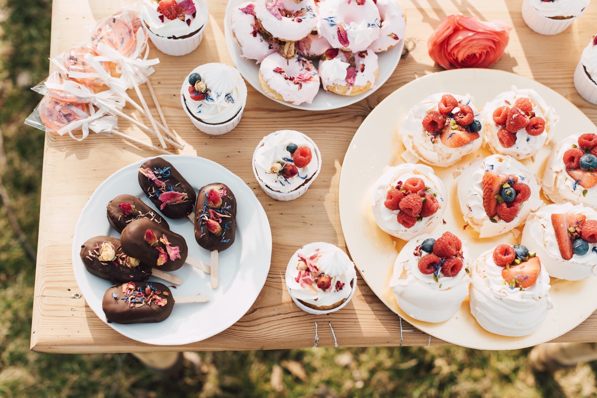 Słodki stół z różnorodnymi deserami: lizaki, babeczki z kremem, donuty z lukrem i mini bezy udekorowane owocami i jadalnymi kwiatami, ustawione na drewnianym stole na zewnątrz.