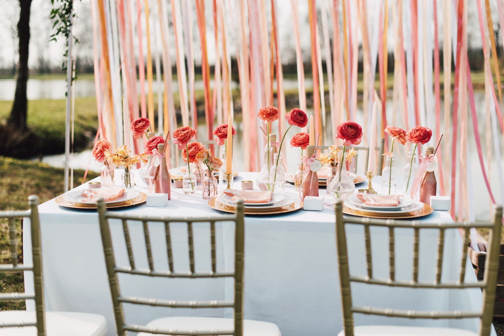 Uroczysty stół na zewnątrz z dekoracjami w odcieniach różu i pomarańczy, kwiaty w wazonach, świece i butelki z etykietami, tło z kolorowych wstążek.