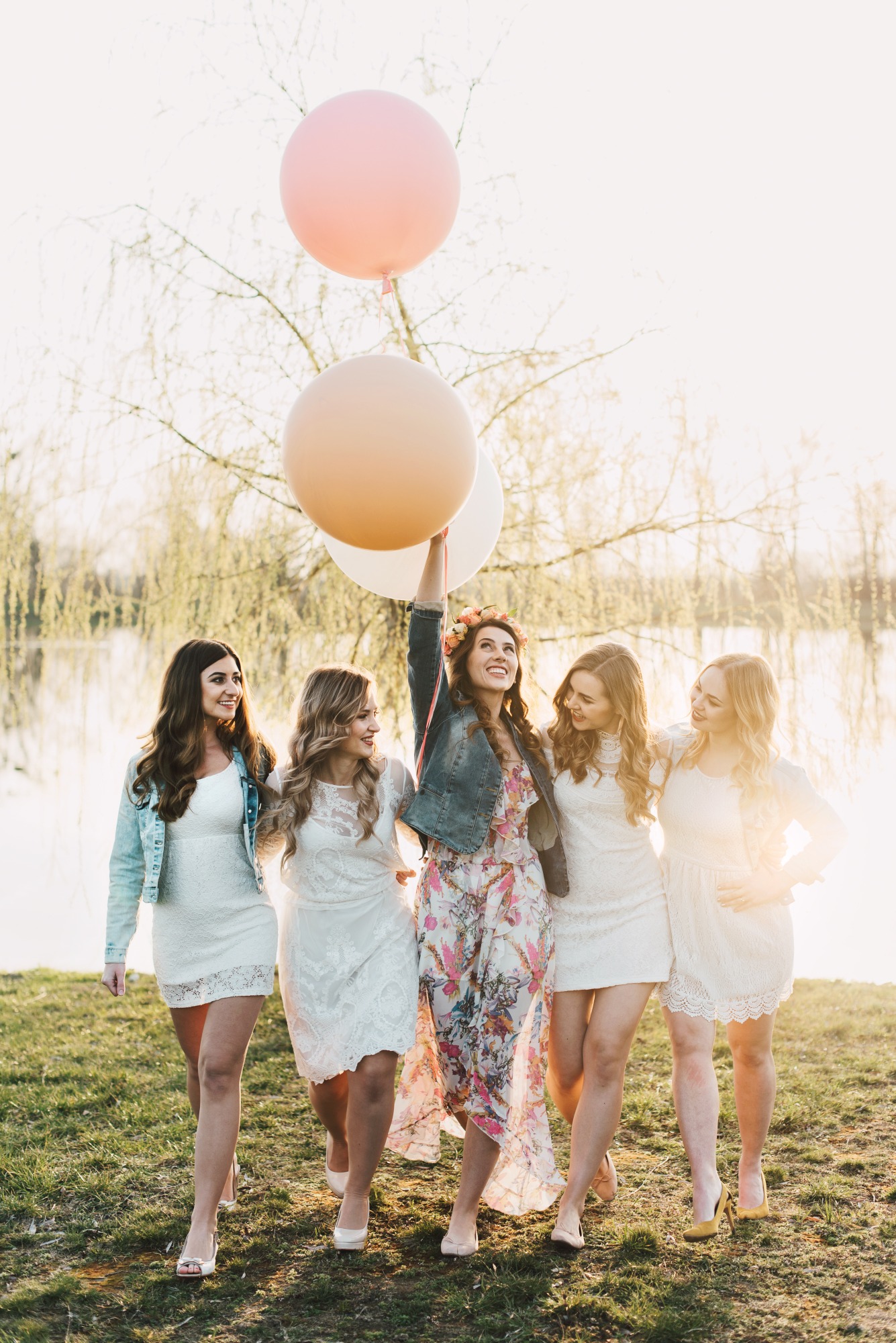 Grupa pięciu uśmiechniętych kobiet w jasnych sukienkach spaceruje po trawie, jedna z nich trzyma w górze trzy pastelowe balony, w tle jezioro i drzewa.
