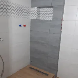 Ukończona łazienka z szarymi i białymi płytkami, wzór w zygzaki na górze, drewnopodobna podłoga z odpływem liniowym, widoczne przyłącza wodne.