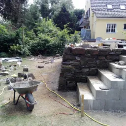 Budowa schodów zewnętrznych z kamienia i betonu, widoczne narzędzia i materiały budowlane, trawa, drzewa i budynek w tle.