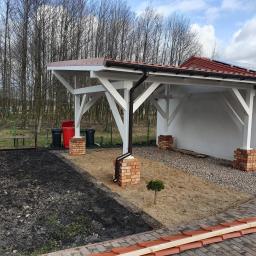 ArtWod - Biała konstrukcja z brązowym dachem, wsparta na ceglanych filarach, z widocznymi panelami słonecznymi na dachu budynku w tle, teren przygotowany pod nasadzenia.