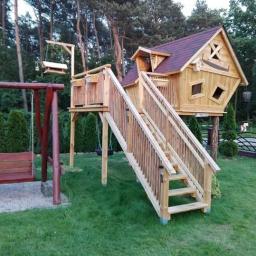 ArtWod - Drewniany domek na drzewie z ukośnymi oknami i schodami, obok huśtawka na tle zielonego ogrodu i drzew.