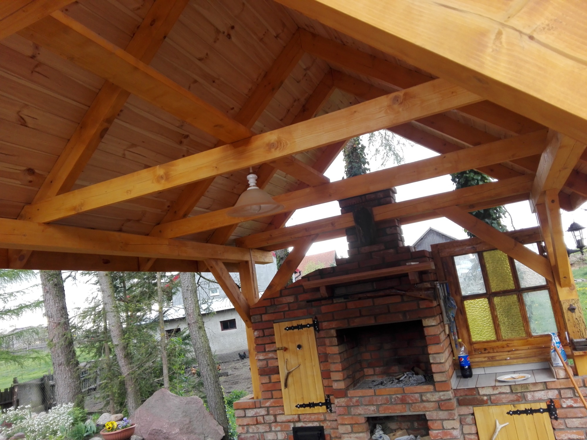 Drewniana konstrukcja zadaszenia z widocznym kominkiem z cegły, elementami dekoracyjnymi i oświetleniem, otoczona zielenią.