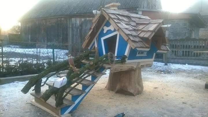Nietypowy domek dla ptaków w kolorze niebieskim, z drewnianym dachem, schodami z gałęzi i imitacją pnia drzewa jako podstawy, umieszczony na zewnątrz w scenerii przypominającej ogród.