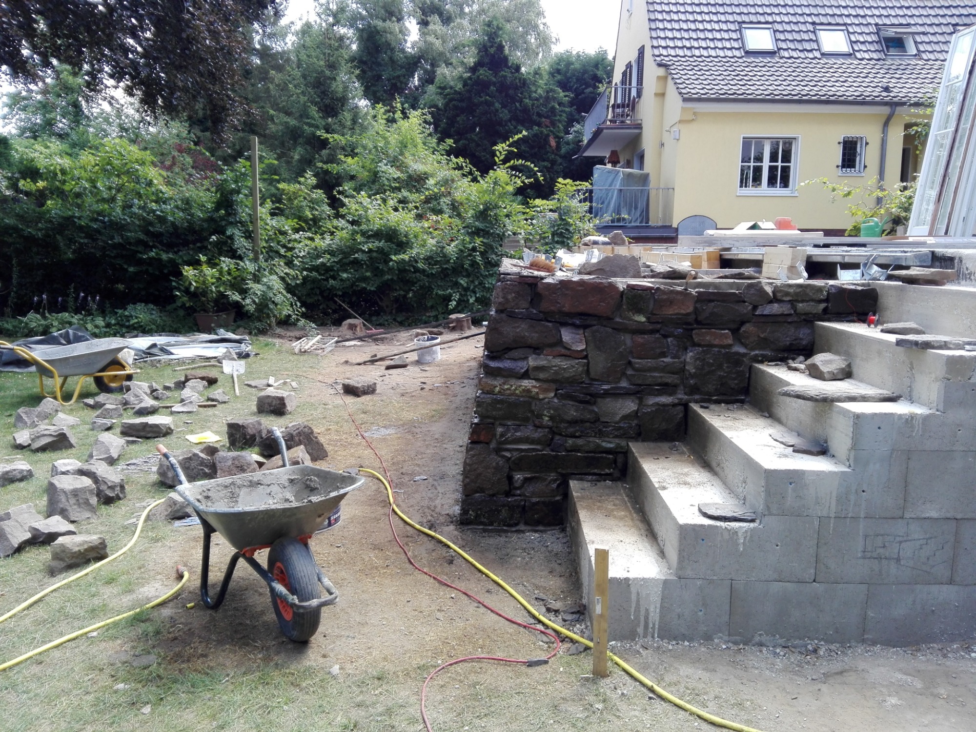 Budowa schodów zewnętrznych z kamienia i betonu, widoczne narzędzia i materiały budowlane, trawa, drzewa i budynek w tle.