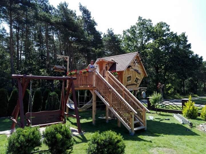 Drewniany domek na drzewie z werandą, schodami, huśtawką i hamakiem w ogrodzie, widok z bliska na tle drzew.