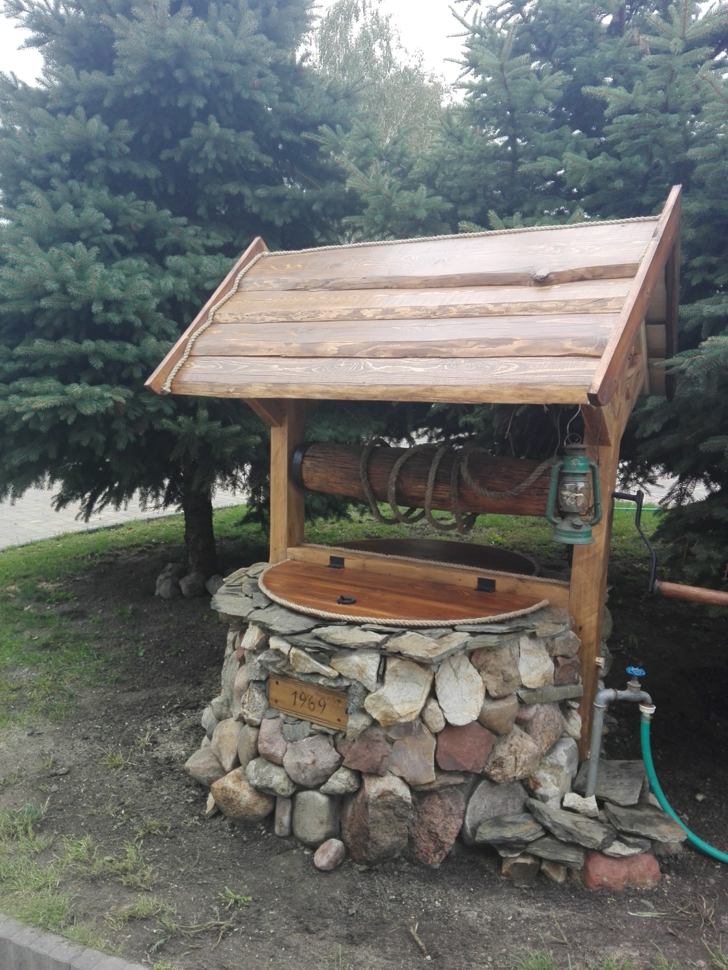 Studnia ogrodowa z drewnianą konstrukcją, kamienną obudową z tabliczką '1969', zieloną latarnią i kranem z wężem, na tle iglaków.