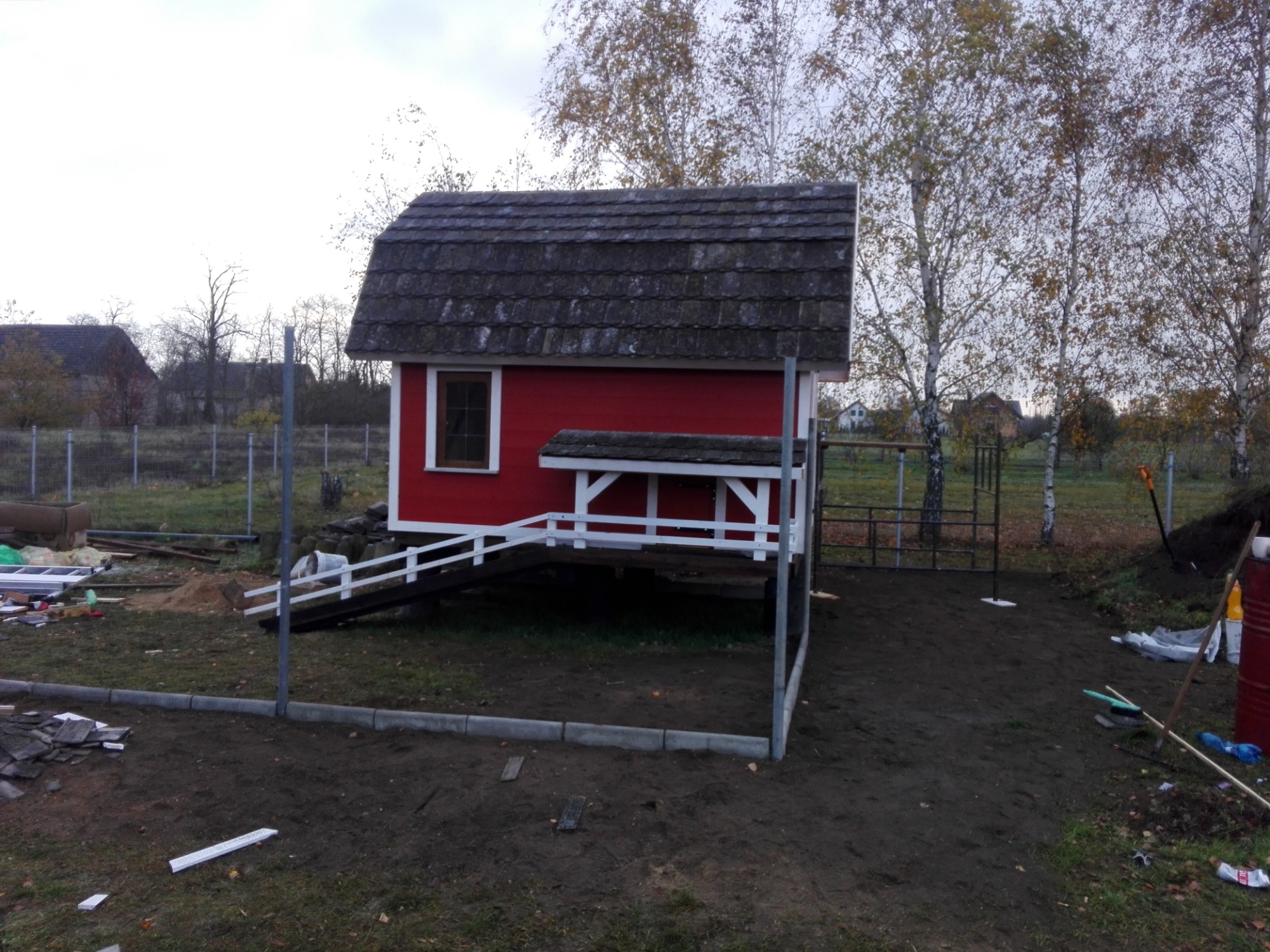 Czerwona, drewniana konstrukcja z ciemnym, spadzistym dachem i białymi detalami, posadowiona na podwyższeniu z rampą, otoczona elementami ogrodzenia i narzędziami budowlanymi, sugerująca etap...